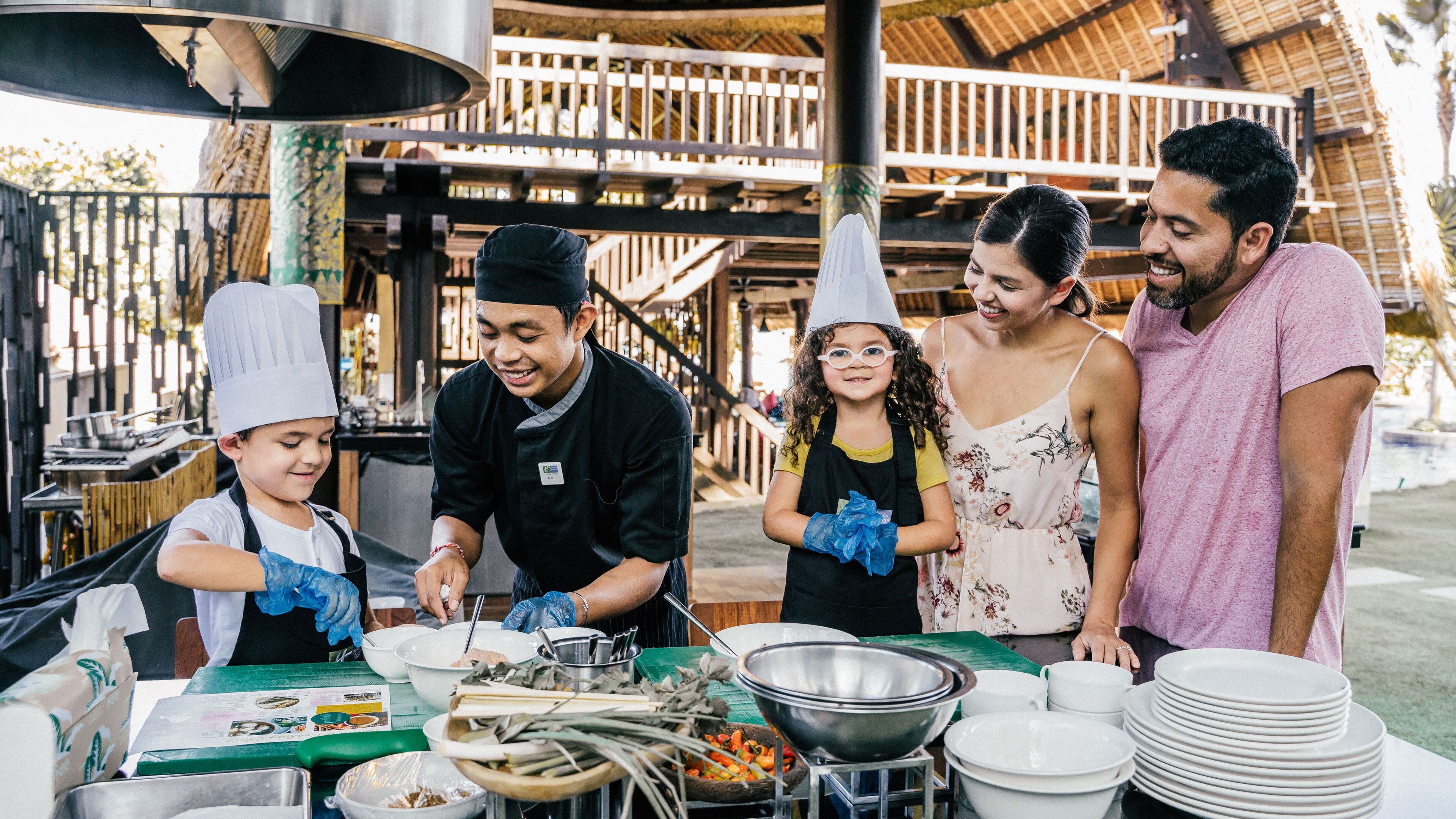 Un grupo de personas preparando comida en una estación de cocina al aire libre, con una terraza de madera y un techo de paja al fondo.