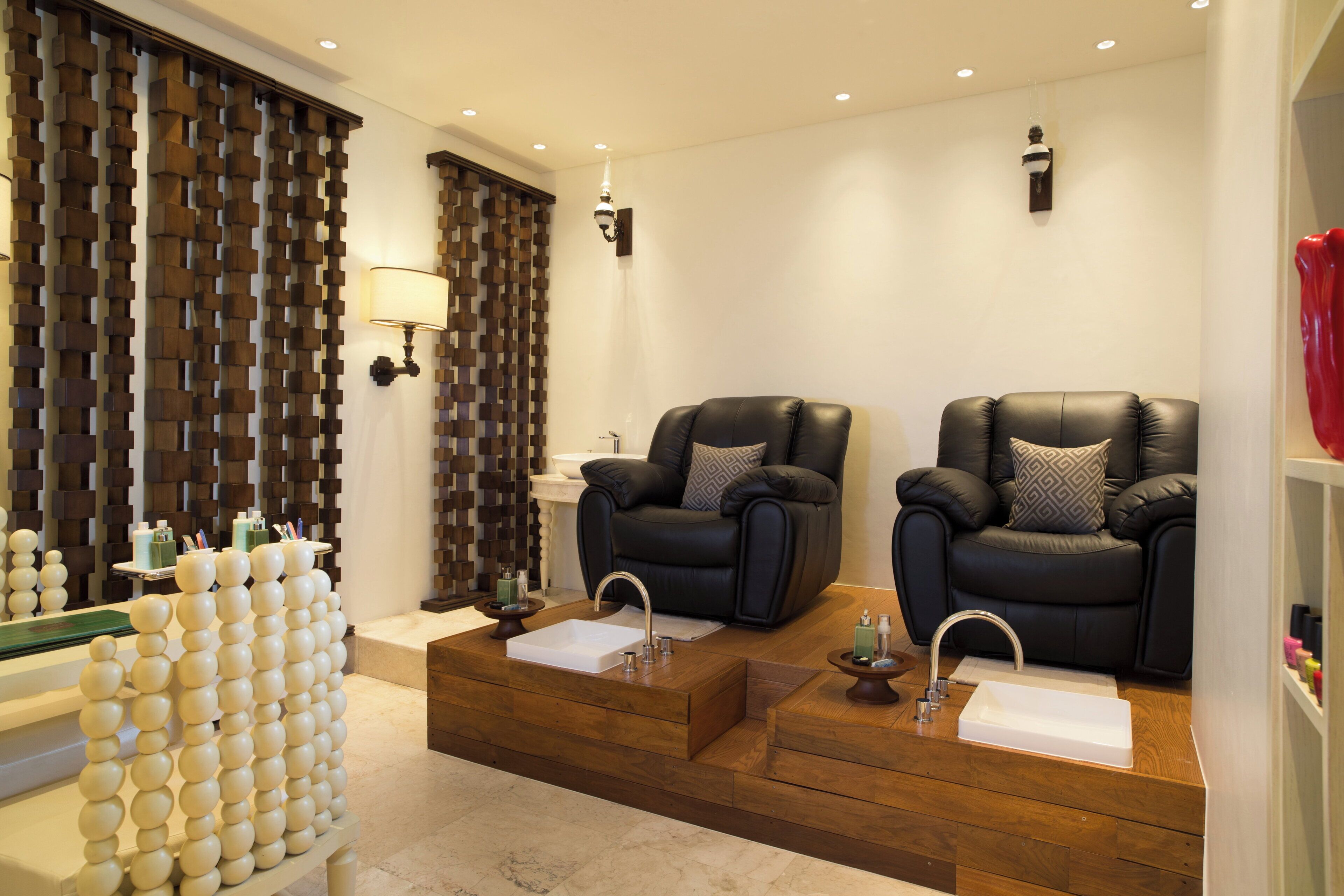 Ein Wellnessraum mit zwei Massagesesseln, einer Holzwand mit geometrischen Mustern, einem weißen Waschbecken und einem kleinen Tisch mit Pflegeprodukten.