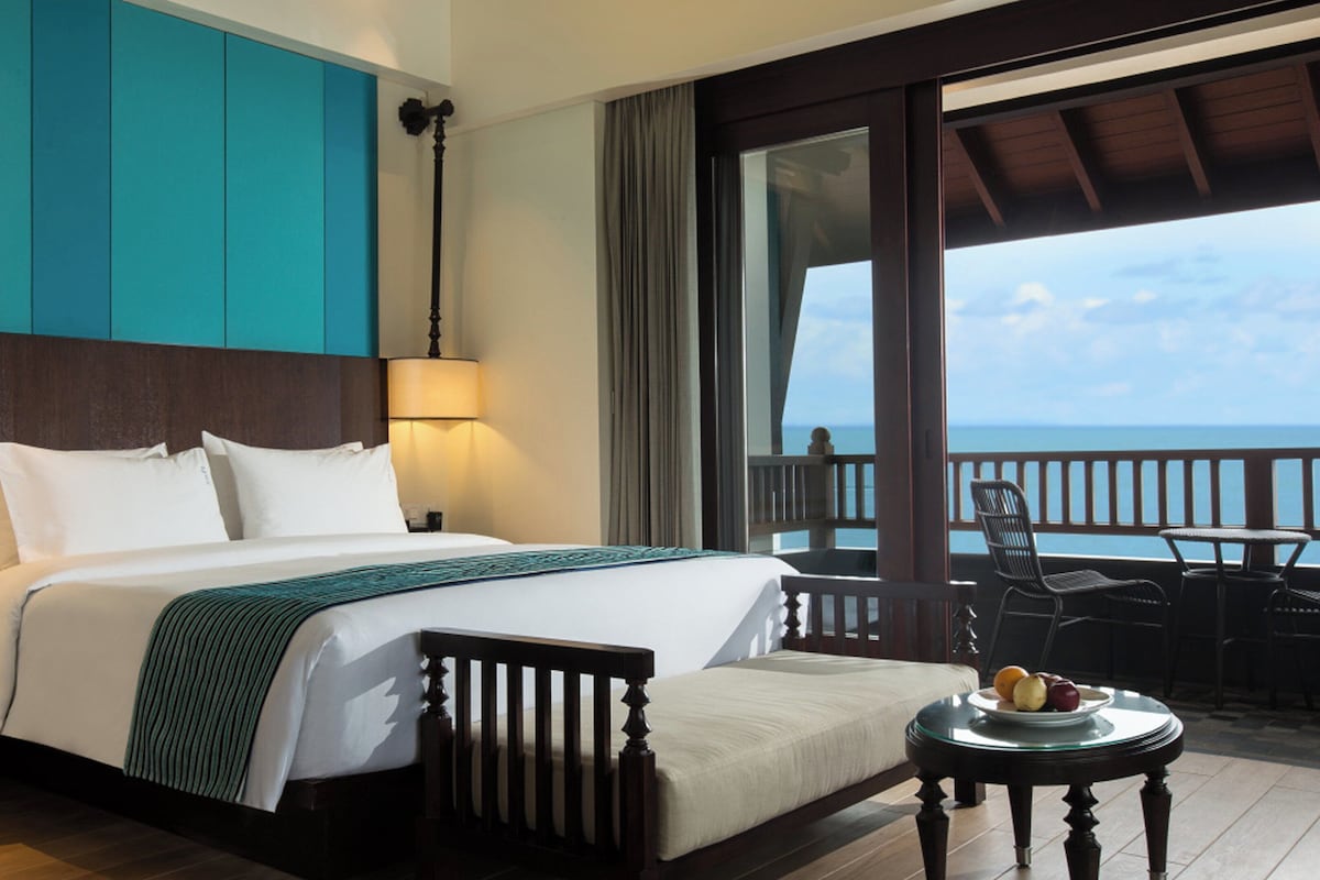 A hotel room with a bed, a nightstand, a table with fruit, and a view of the ocean.