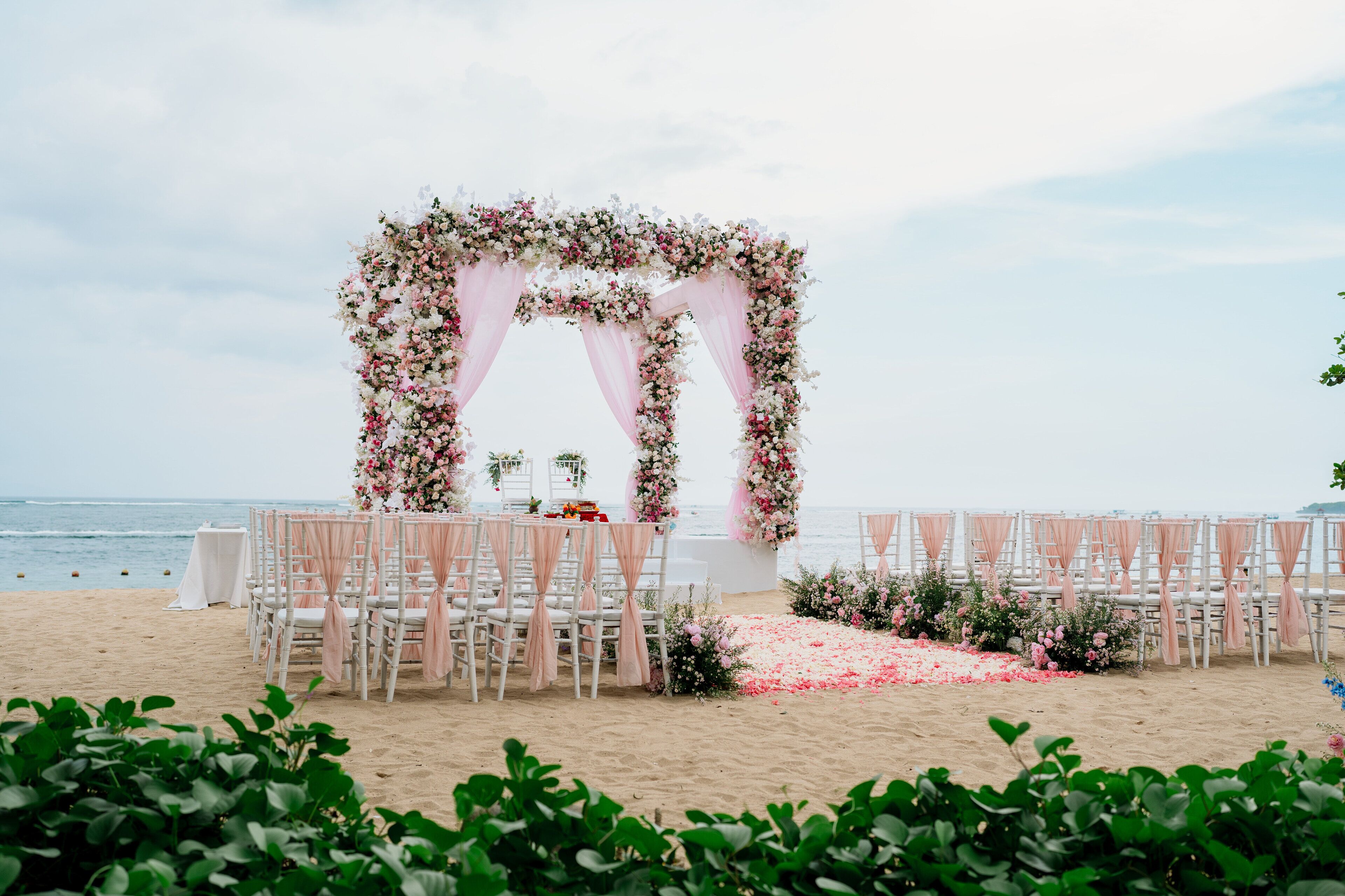 A beach wedding setup with a floral arch, white chairs, and a white table.