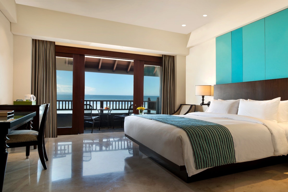 A hotel room with a bed, desk, and chairs near a balcony with a view of the ocean.