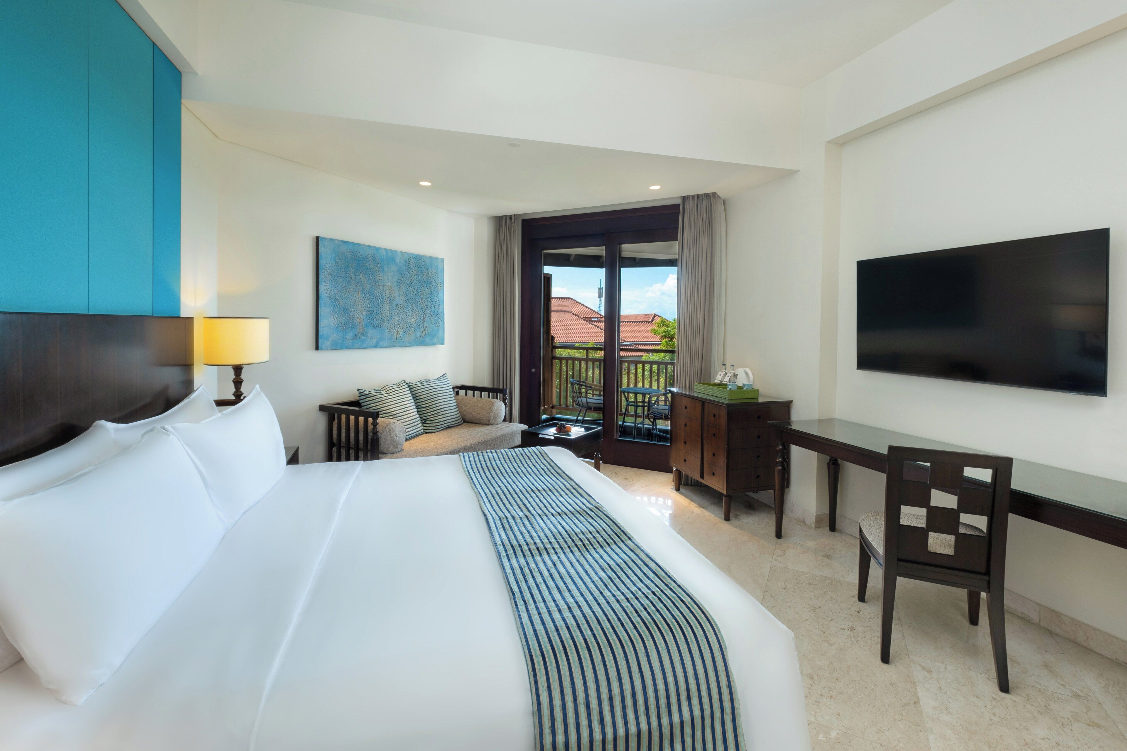 A hotel room with a large bed, a desk, a chair, a TV, a balcony with a view, and a painting on the wall.