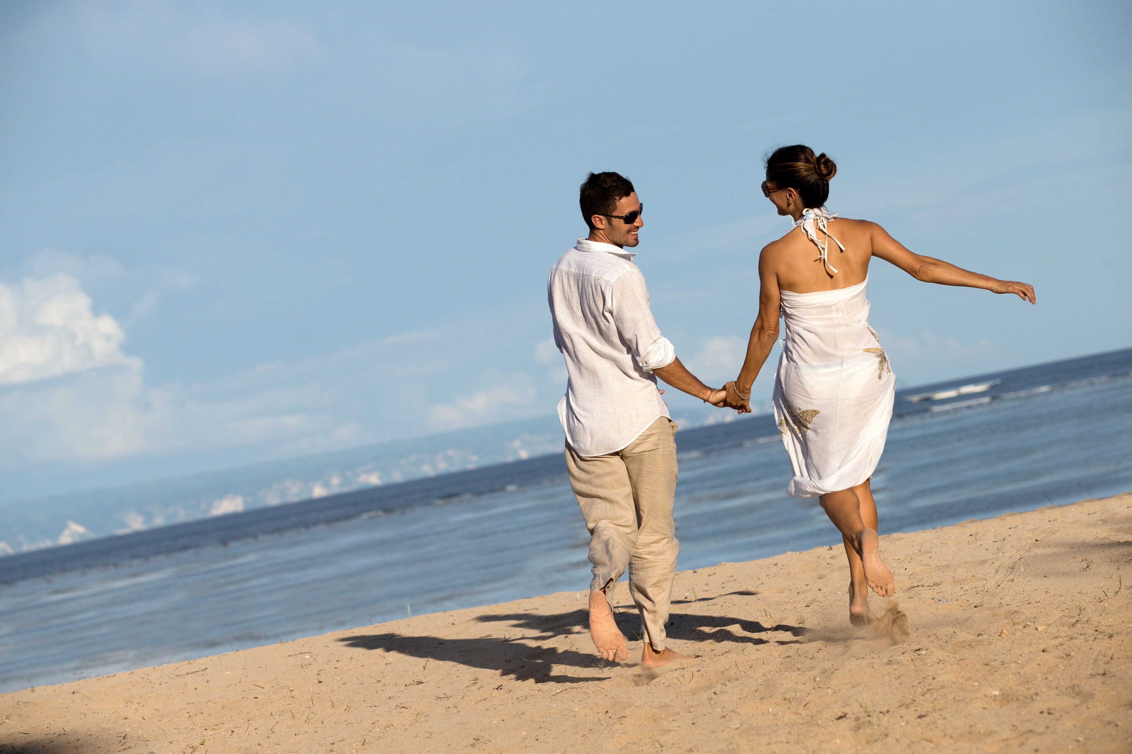 A couple holding hands on a sandy beach with the ocean in the background.