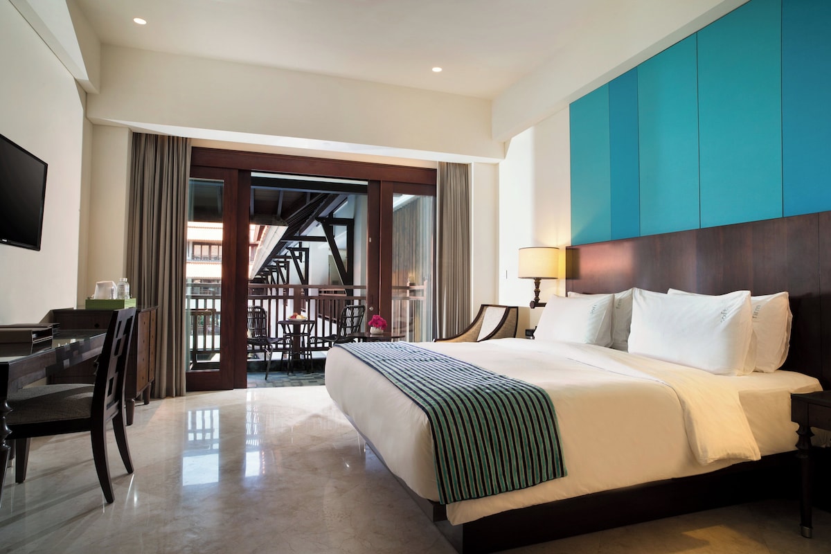 A hotel room with a large bed, a desk, a chair, a TV, and a balcony with a view.