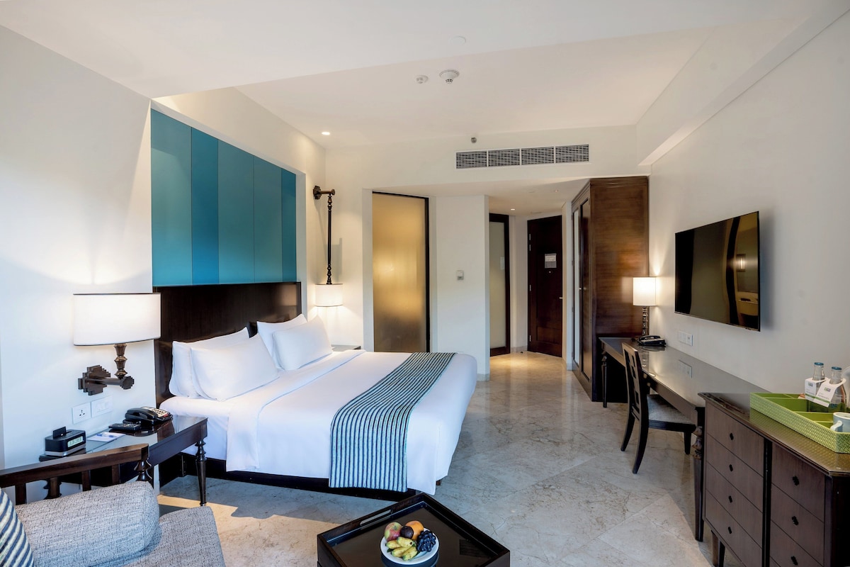 A modern hotel room with a large bed, a desk, a chair, a coffee table with fruit, and a TV.