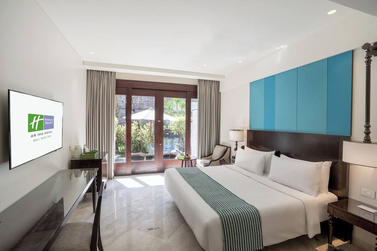 A hotel room with a large bed, a TV on the wall, a desk, and a view of the outdoors.
