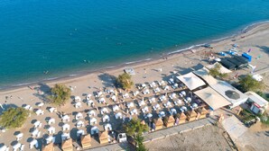 An aerial view of a beach with numerous white umbrellas and sun loungers arranged in rows, a few thatched-roof huts, and a clear view of the sea. - Unluselek Hotel (Anamur)