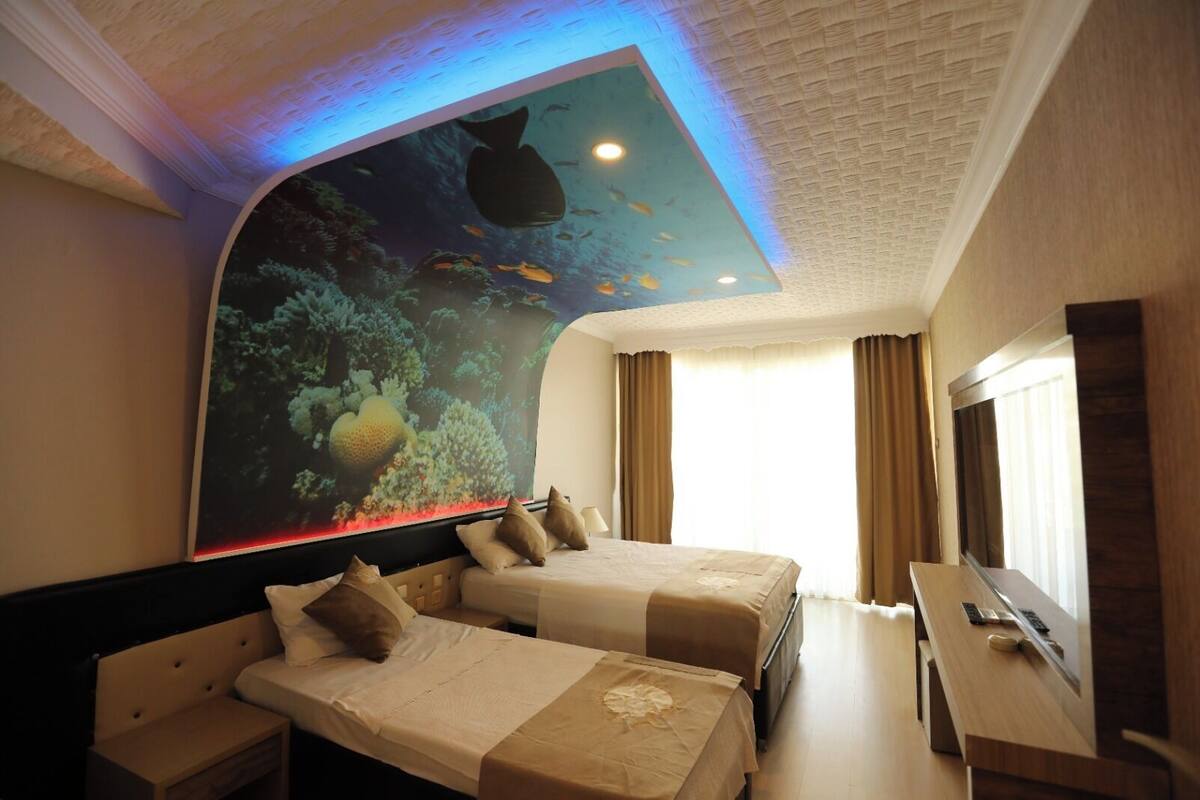 Quarto de hotel com duas camas, um mural grande na parede retratando uma cena subaquática, uma TV de tela plana e uma escrivaninha com computador.