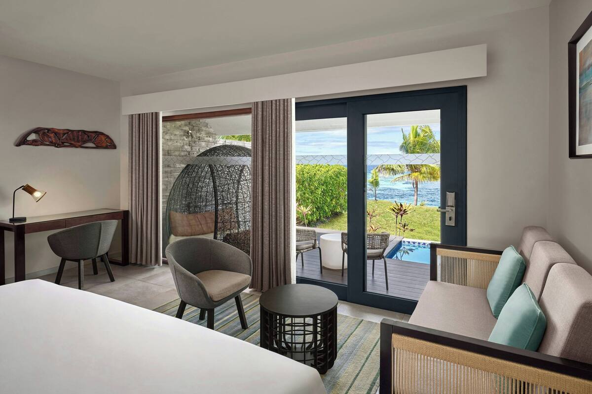 A hotel room with a bed, a desk, a chair, a sofa, a small table, and a view of a pool and ocean.
