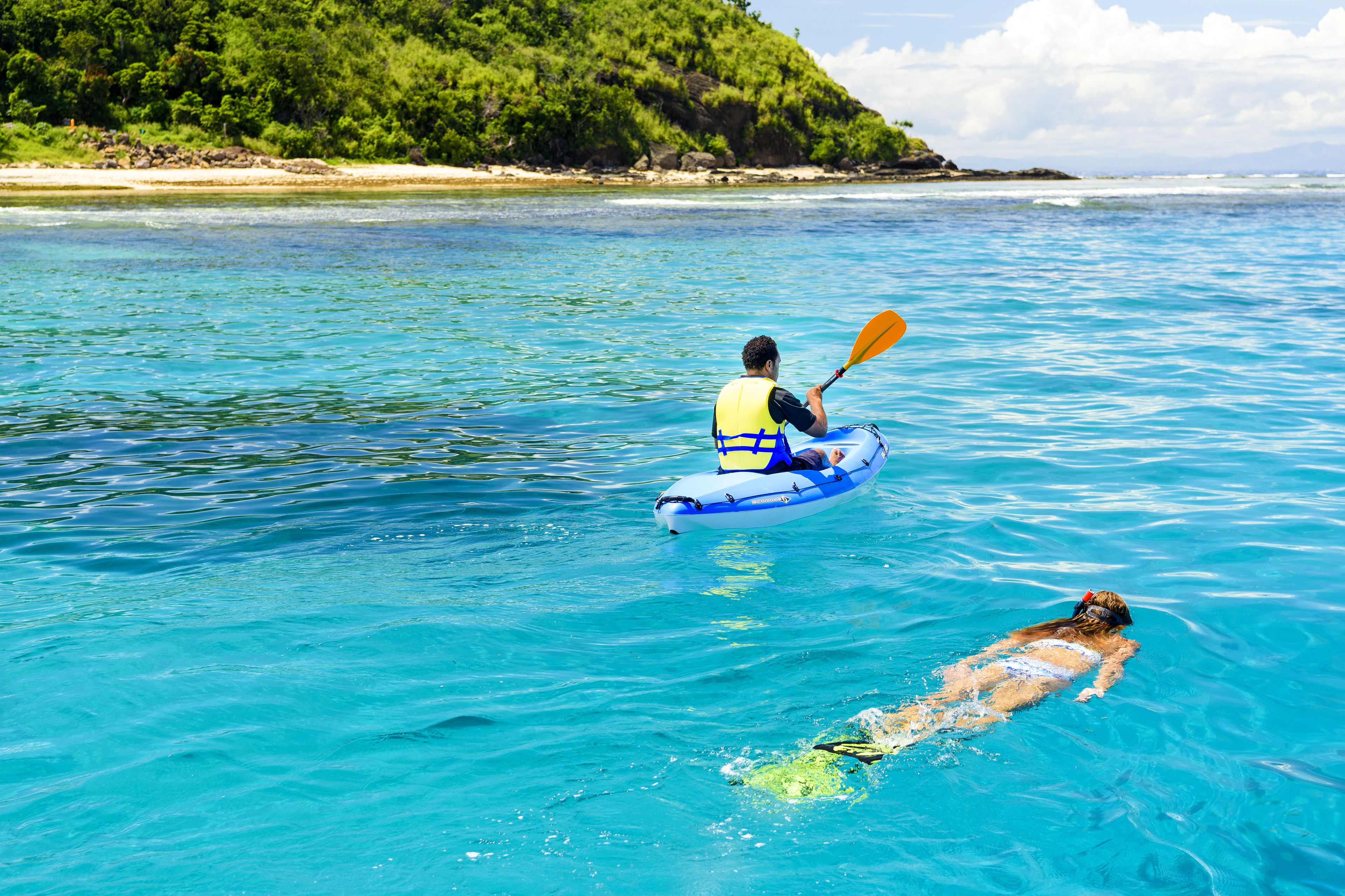 A person kayaking on a clear blue sea with a forested island in the background.