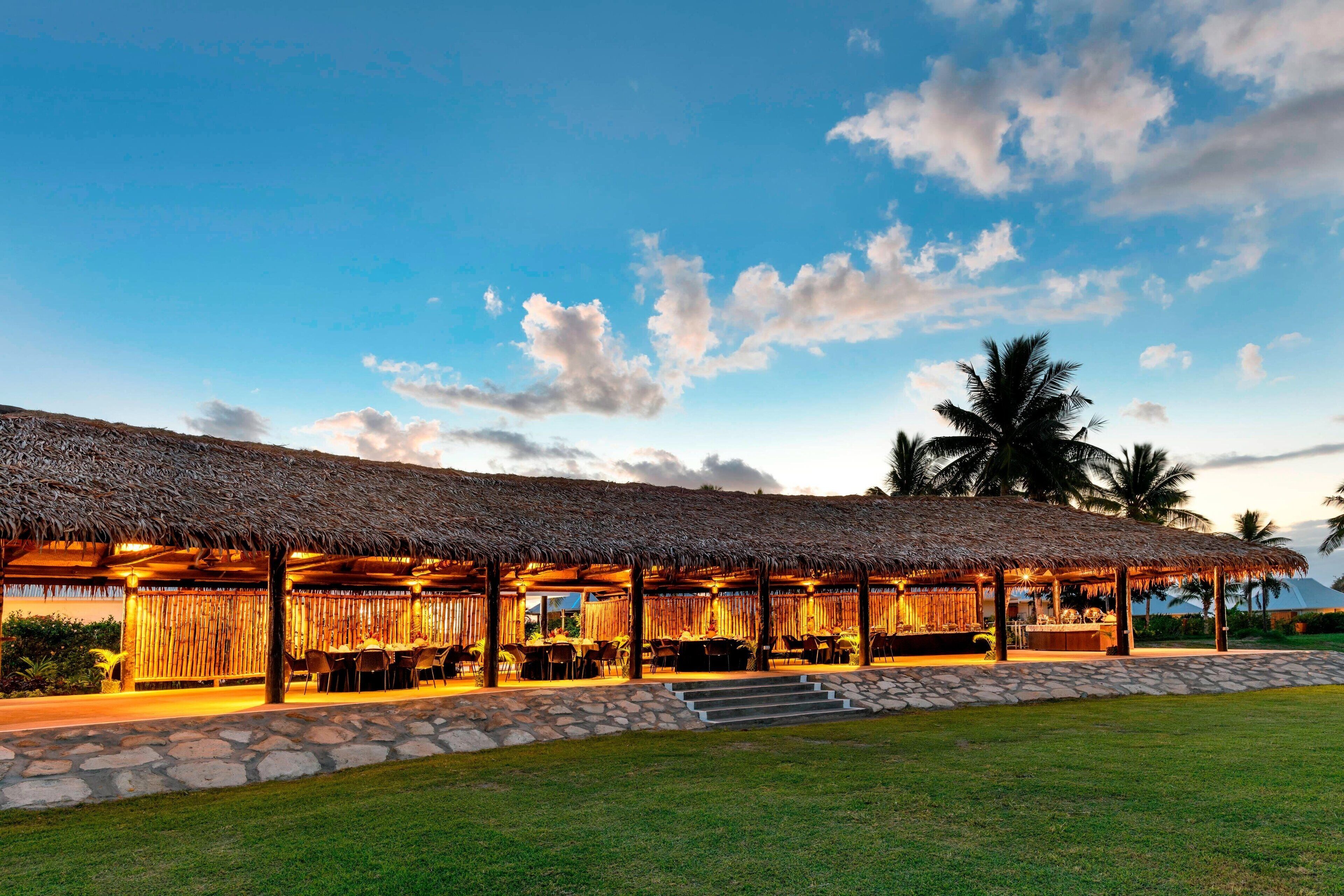 A thatched-roof outdoor dining area with stone steps leading up to it, surrounded by a well-maintained lawn.