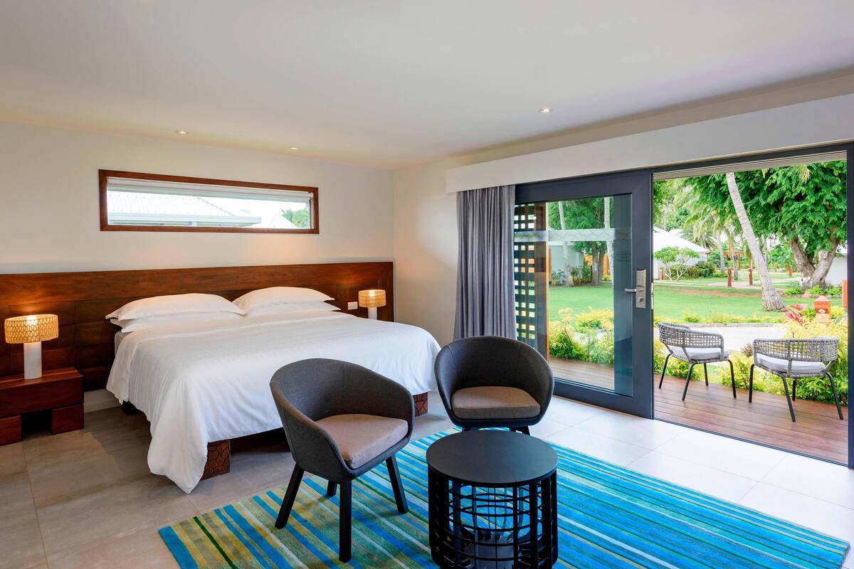 A modern hotel room with a large bed, two armchairs, a small table, and a view of a garden through a glass door.