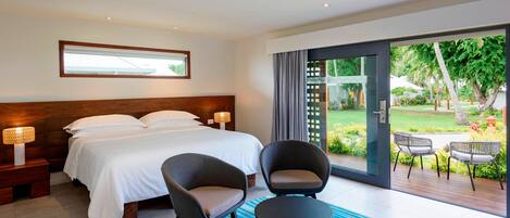 A modern hotel room with a large bed, two armchairs, a small table, and a view of a garden through a glass door.
