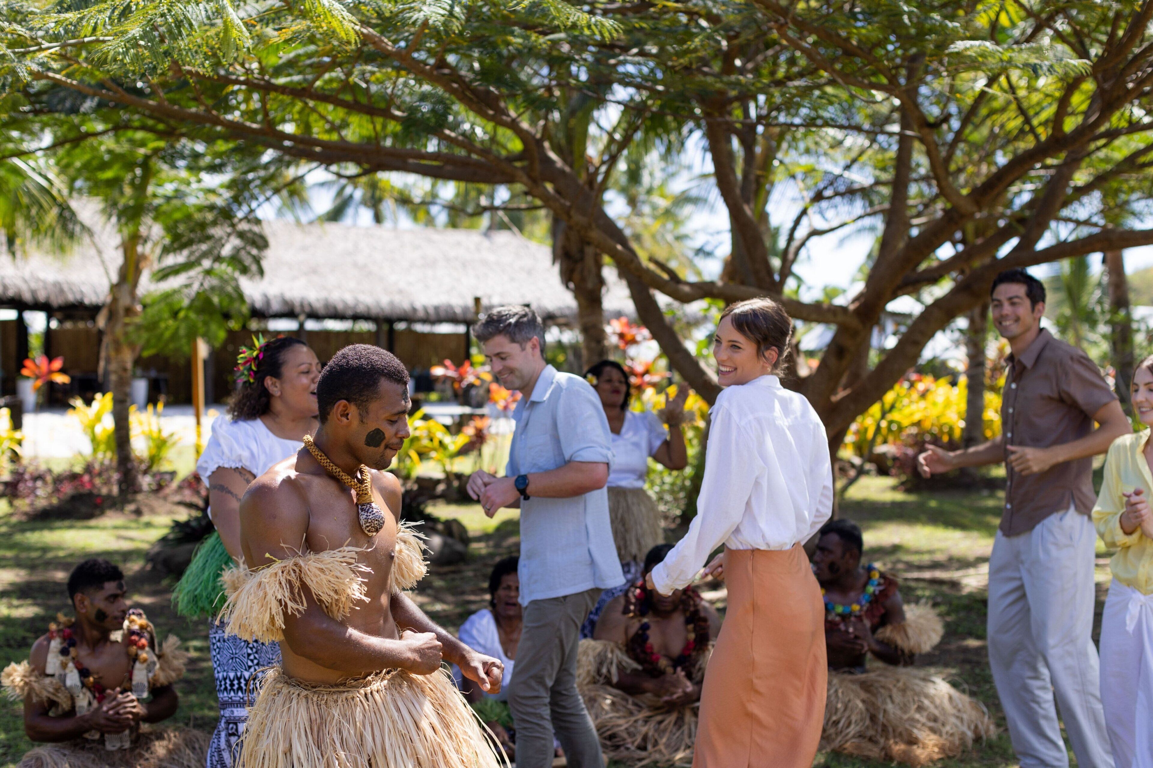 A tropical outdoor setting with people in traditional attire and casual clothing, surrounded by trees and thatched-roof structures.