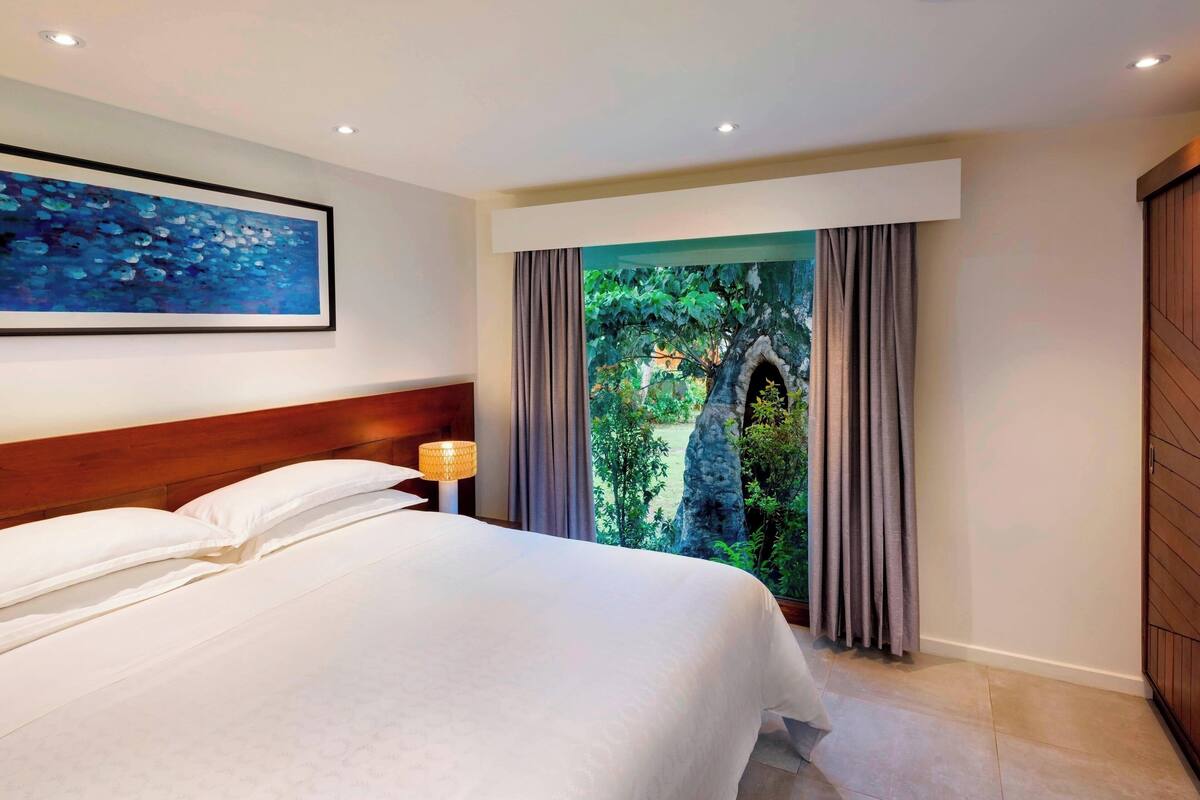 A hotel room with a large bed, a headboard, a nightstand with a lamp, a painting on the wall, and a view of a forest through a window.