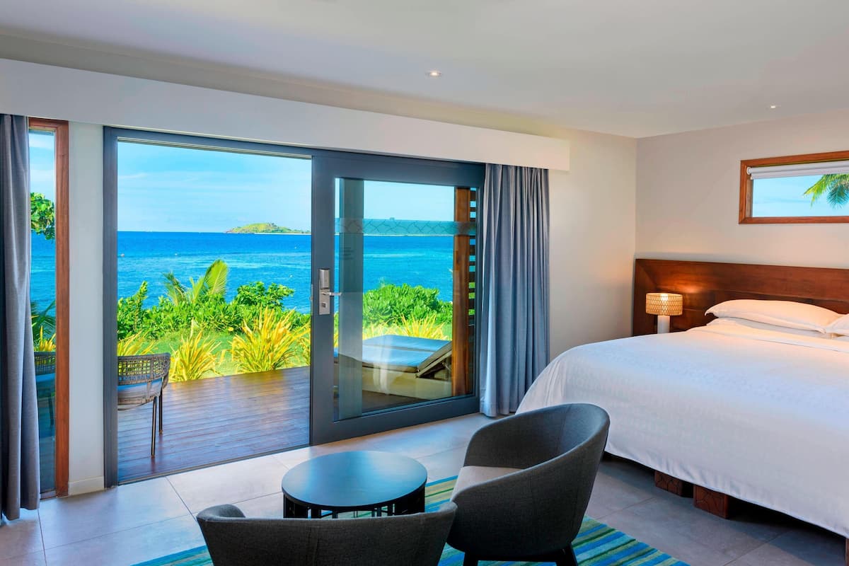 A hotel room with a large bed, a seating area with a table, and a view of the ocean through a sliding glass door.