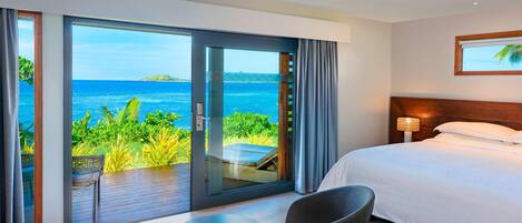 A hotel room with a large bed, a seating area with a table, and a view of the ocean through a sliding glass door.
