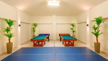 Una sala con dos mesas de ping pong, dos mesas de futbolín y cuatro plantas en macetas.