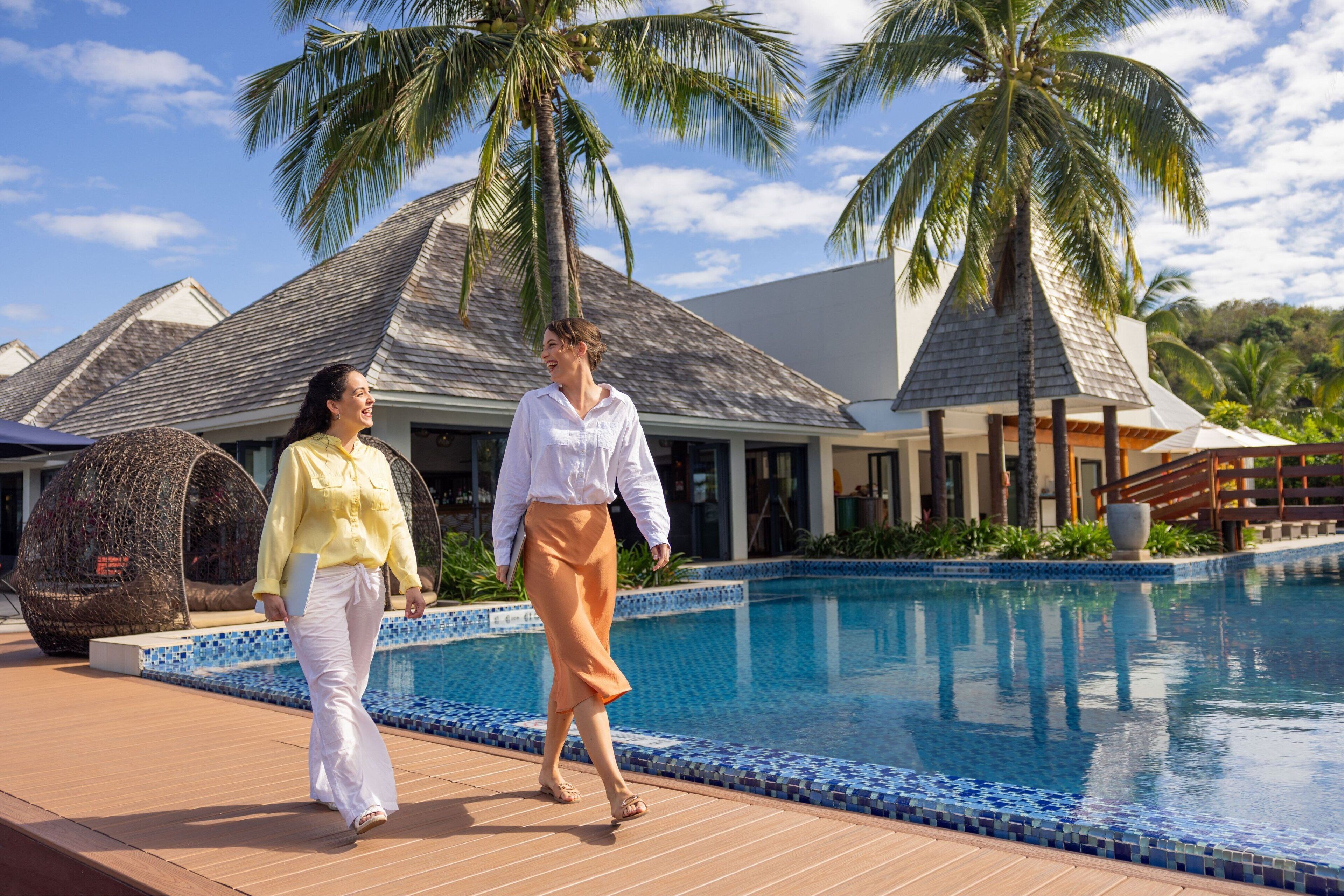 A resort pool area with palm trees, a covered seating area, and a building with a tiled roof.