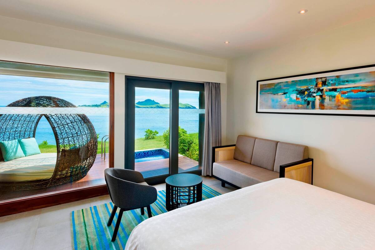 A hotel room with a bed, a sofa, a chair, a small table, and a large window offering a view of the sea and a beach.