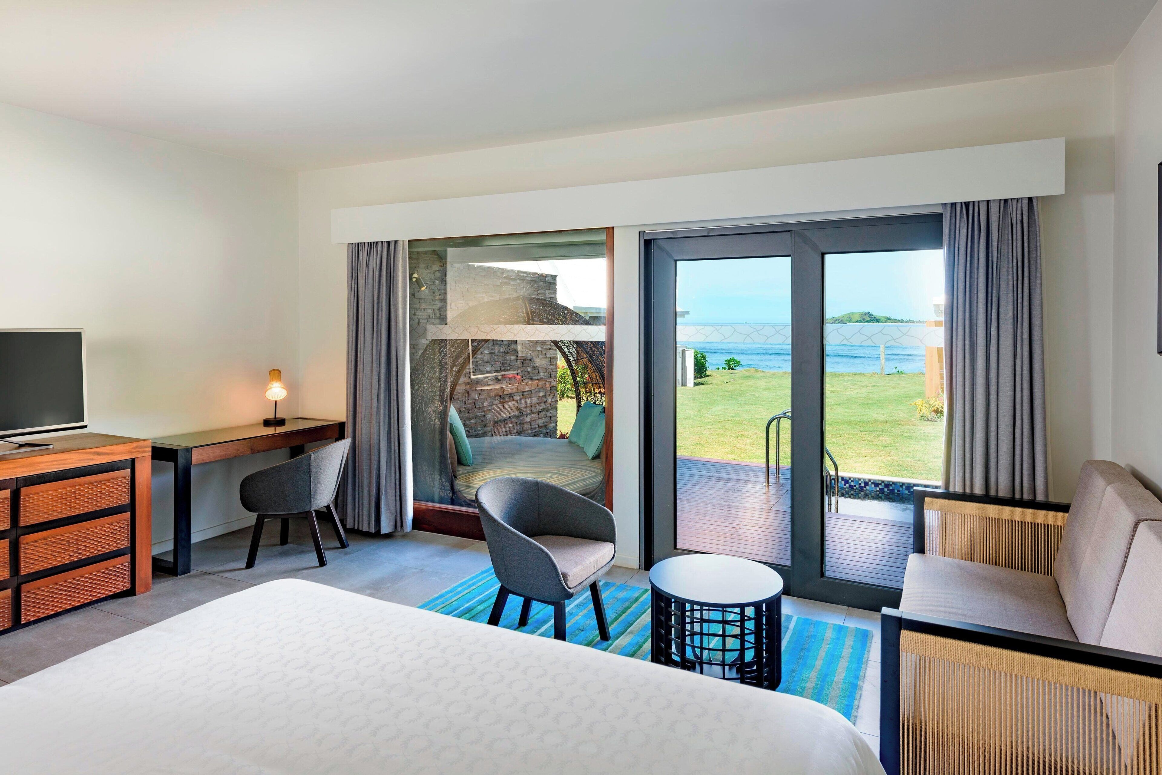 Habitación de hotel con cama, escritorio, silla, una mesita pequeña y un sofá. Cuenta con un ventanal grande con vista al mar y a un área verde.
