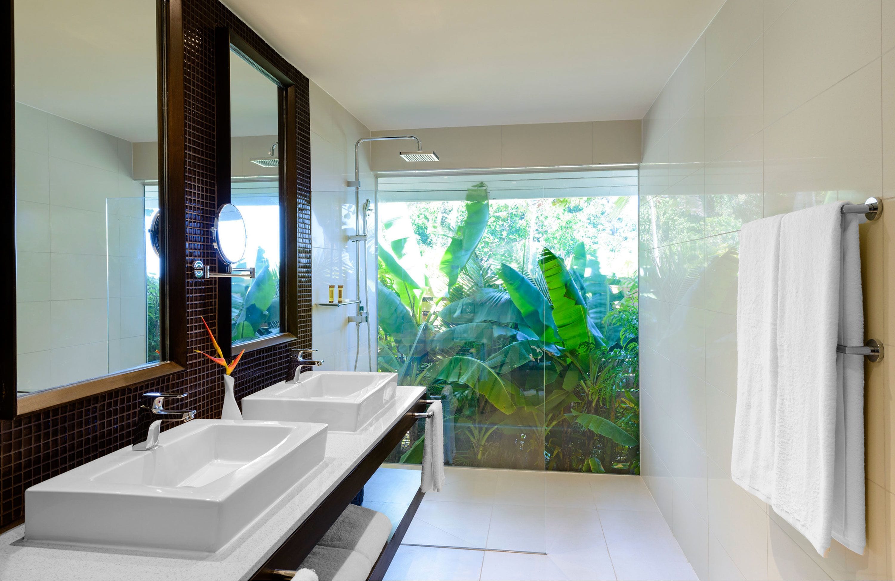 Ein modernes Badezimmer mit zwei Waschbecken, einem großen Spiegel und einer Duschkabine aus Glas mit Blick auf üppige tropische Pflanzen.