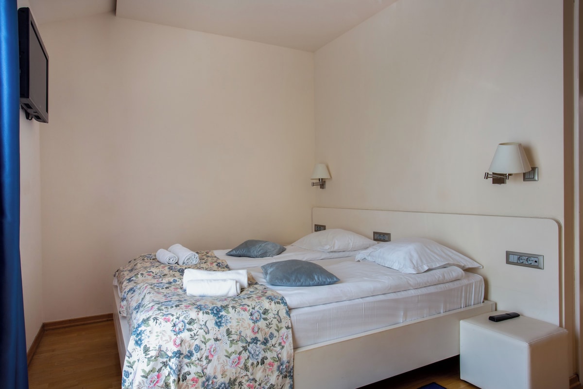 Un dormitorio con una cama, almohadas, una mesita de noche, una lámpara y un televisor instalado en la pared.