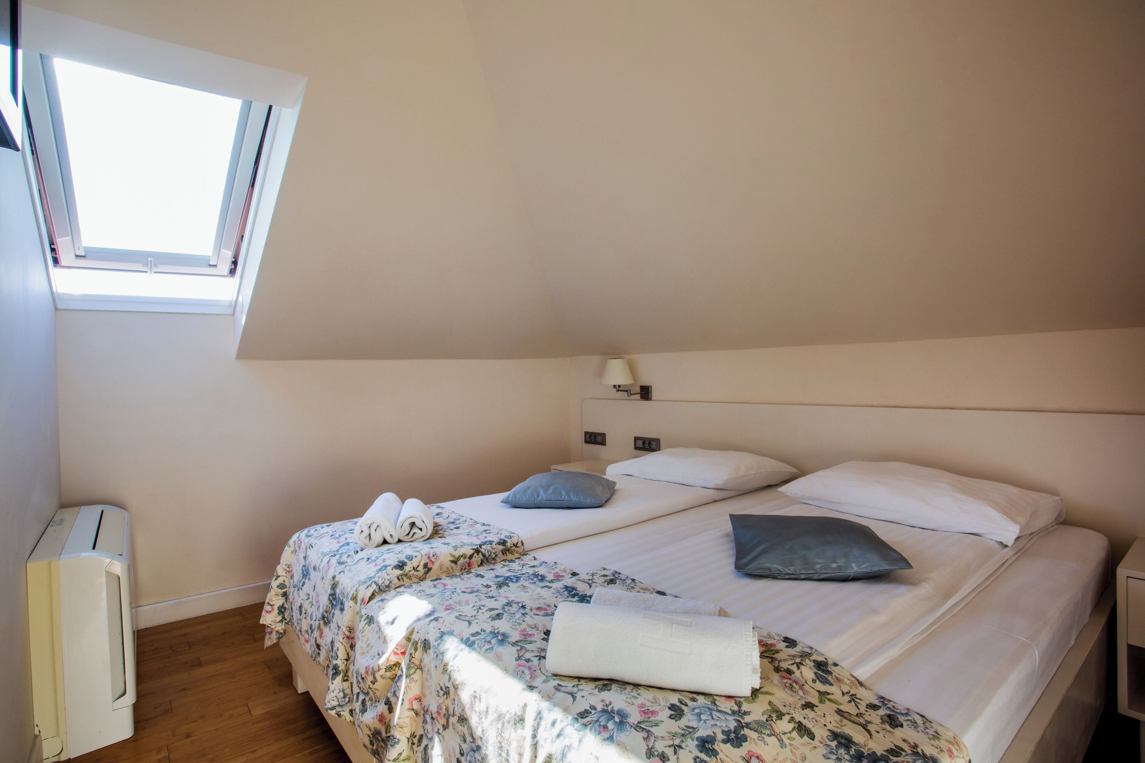 A bedroom with a skylight, a bed with floral bedding, a bedside table with a lamp, and a radiator.