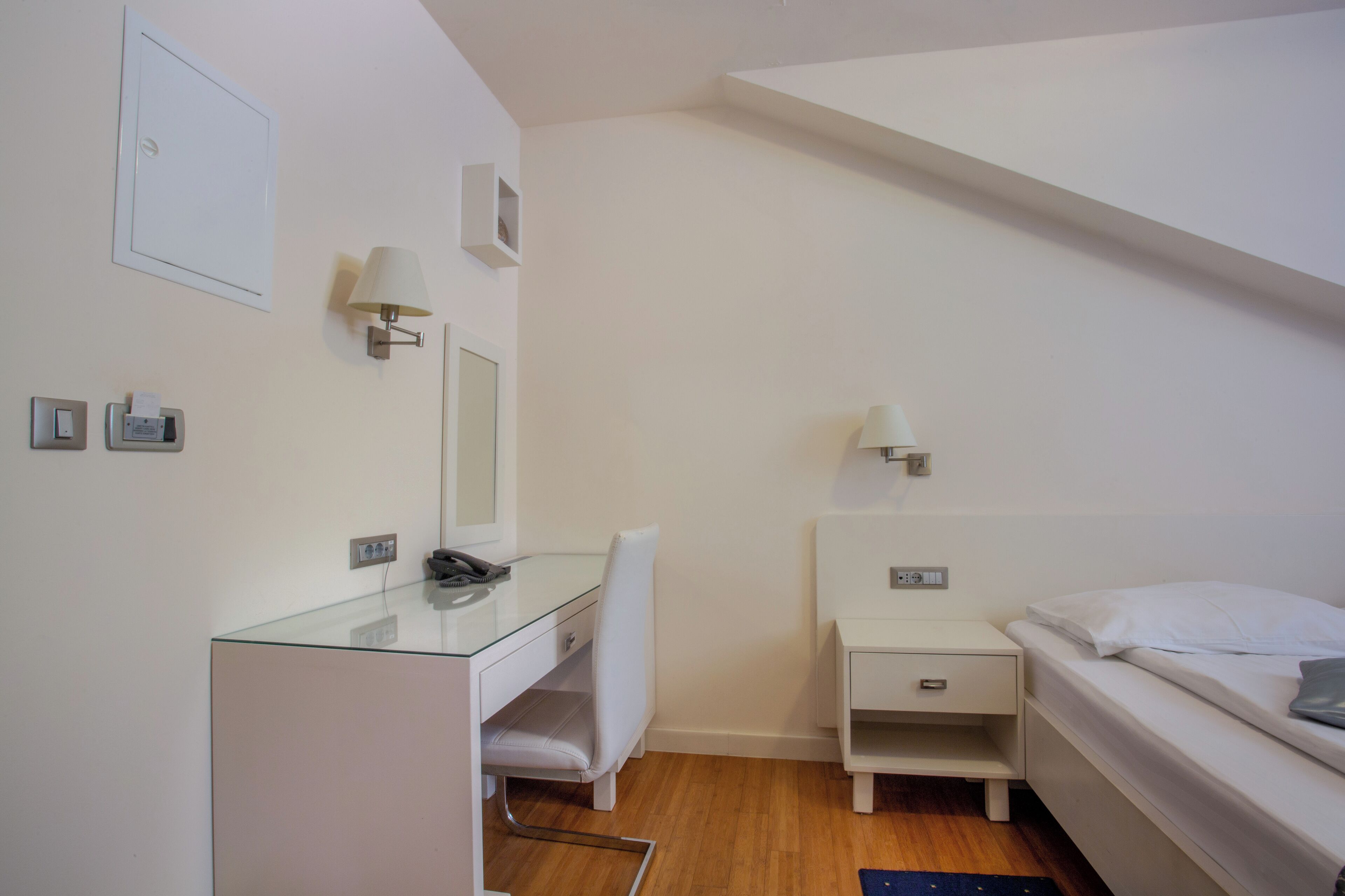 Quarto de hotel moderno com escrivaninha de vidro, uma cadeira branca, uma cama e uma pequena mesa de cabeceira.