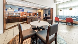 A modern hotel lobby with a round dining table, comfortable seating, a kitchen area, and a large window with patterned curtains.