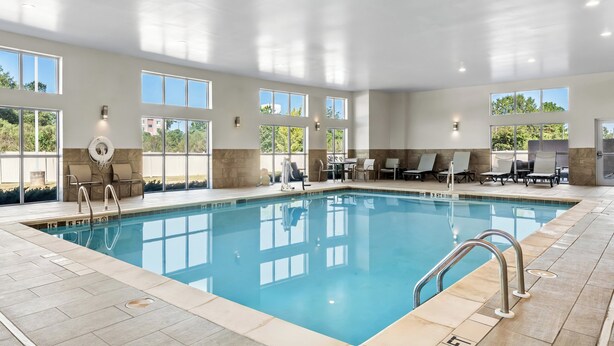 An indoor swimming pool with clear blue water, surrounded by lounge chairs and sunbeds, large windows, and a tiled floor.