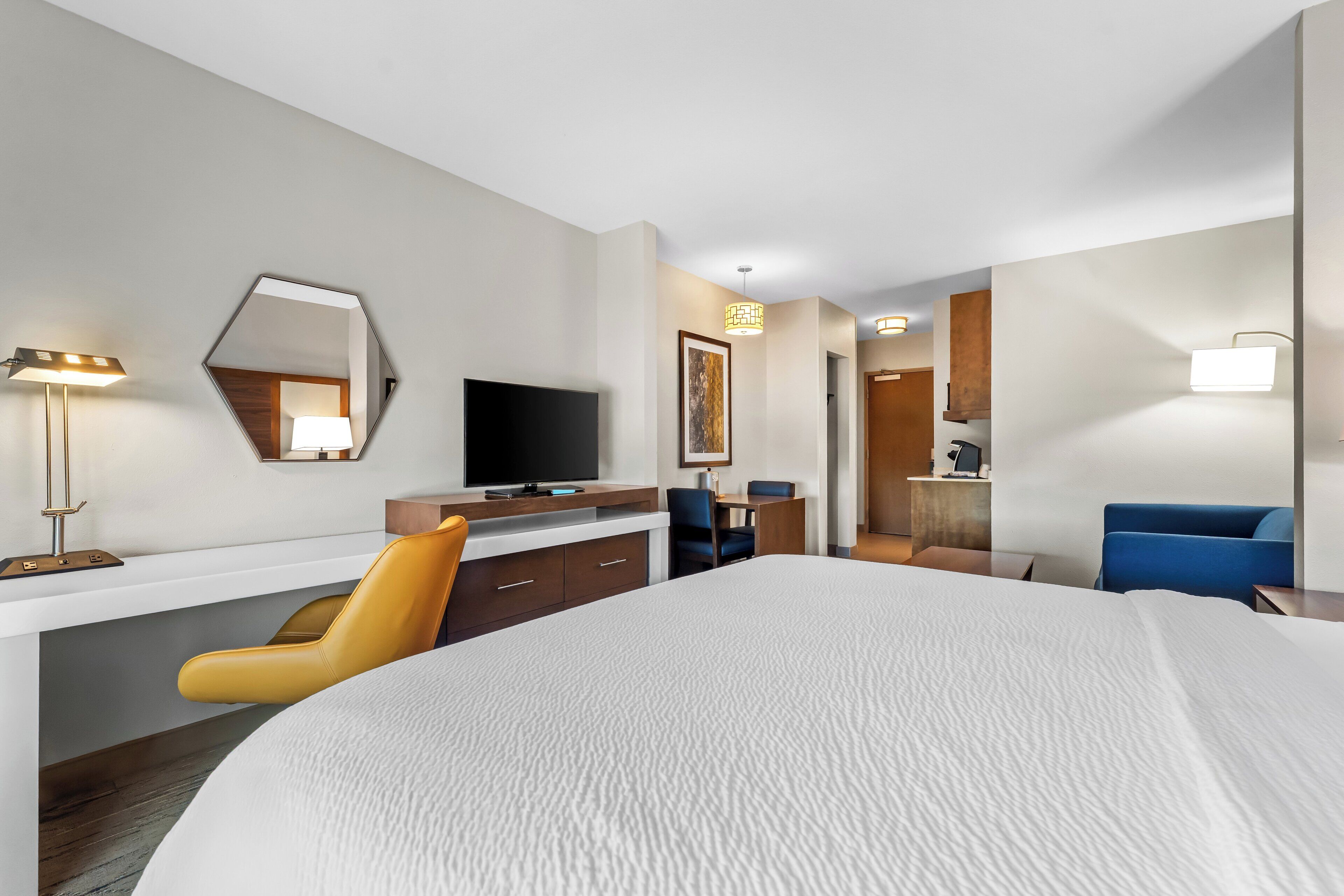 A hotel room with a large bed, a desk with a flat-screen TV, a chair, a desk lamp, and a mirror.