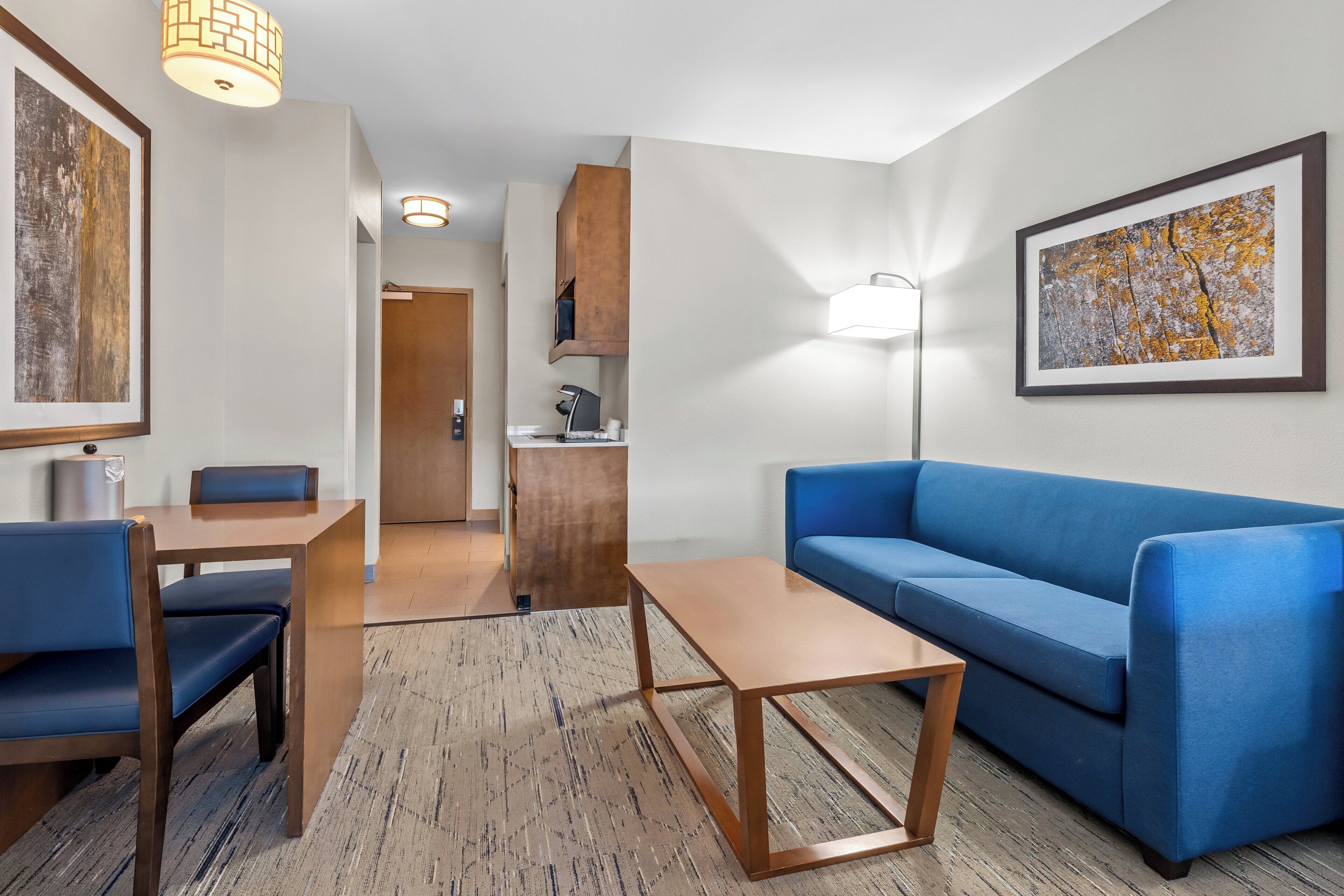A modern hotel room with a blue sofa, wooden coffee table, and a dining area with a table and chairs.