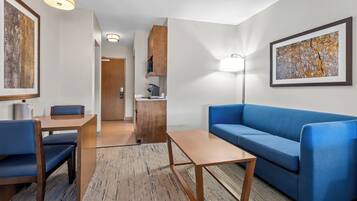 A modern hotel room with a blue sofa, wooden coffee table, and a dining area with a table and chairs.
