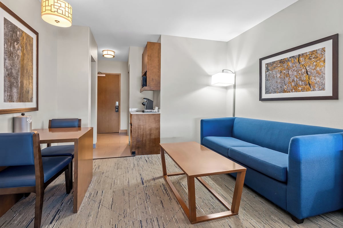A modern hotel room with a blue sofa, wooden coffee table, and a dining area with a table and chairs.