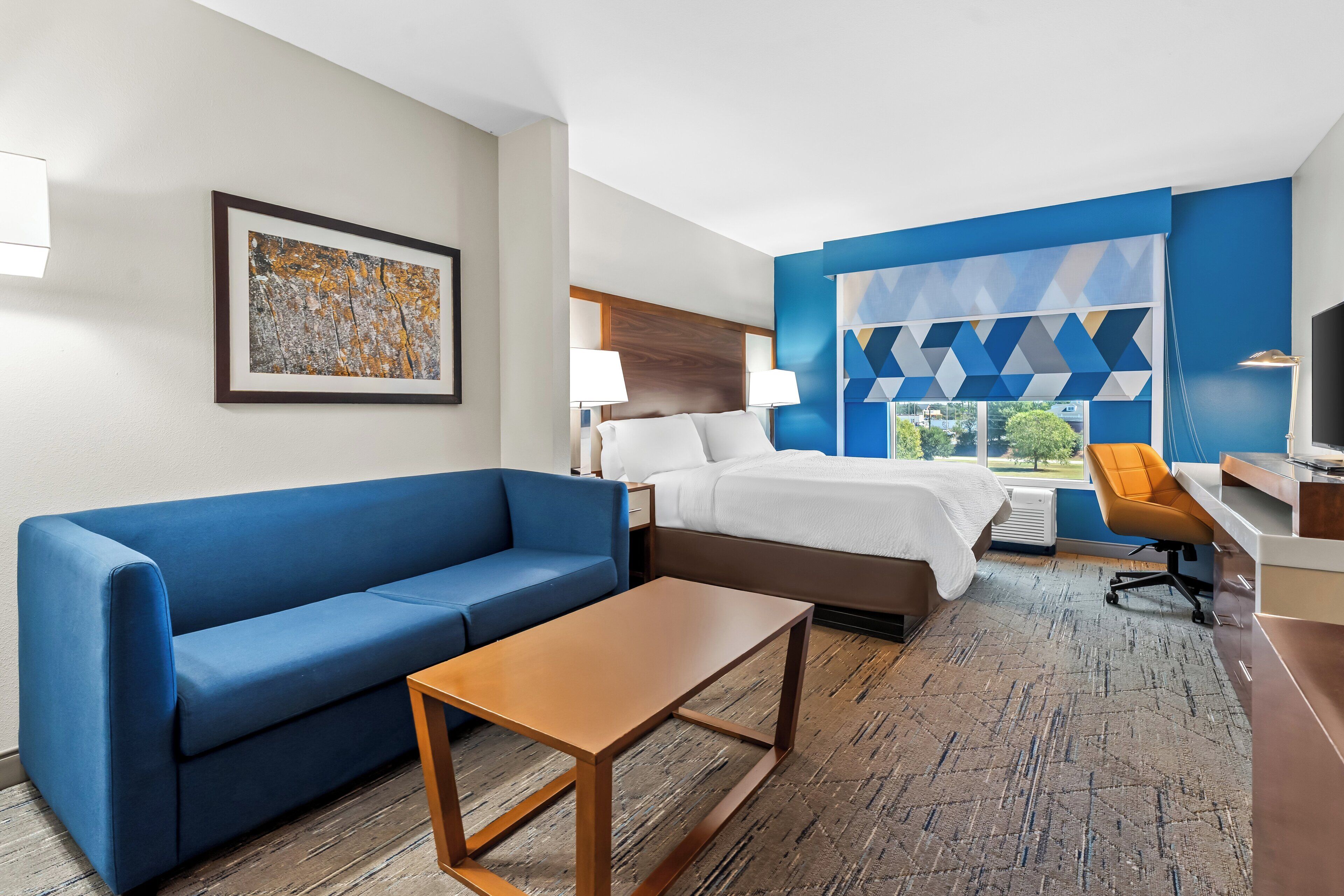 A hotel room with a blue sofa, a bed with white bedding, a wooden coffee table, and a large window with a geometric view.