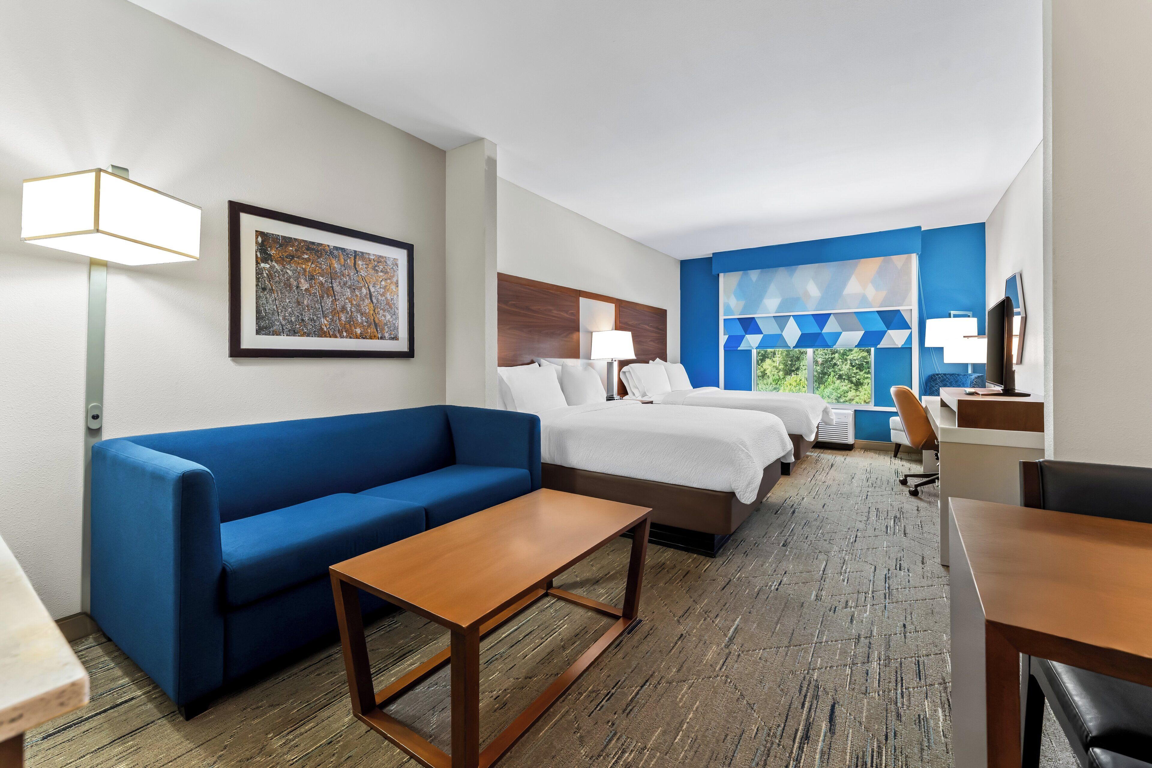 A hotel room with a blue sofa, a wooden coffee table, a bed with white linens, a desk with a chair, and a large window with blue geometric curtains.
