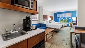 A hotel room with a kitchenette, a bed, a blue sofa, a desk, and a view of greenery.