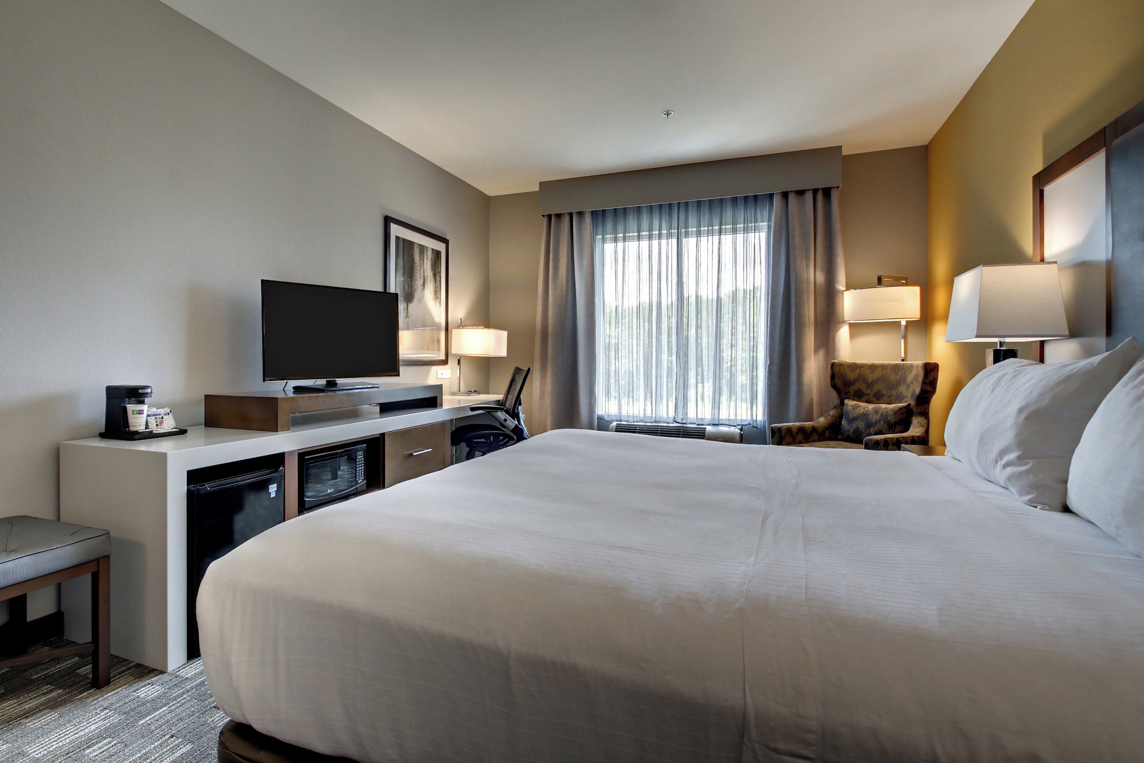 A hotel room with a large bed, a desk with a television, a chair, and a window with curtains.