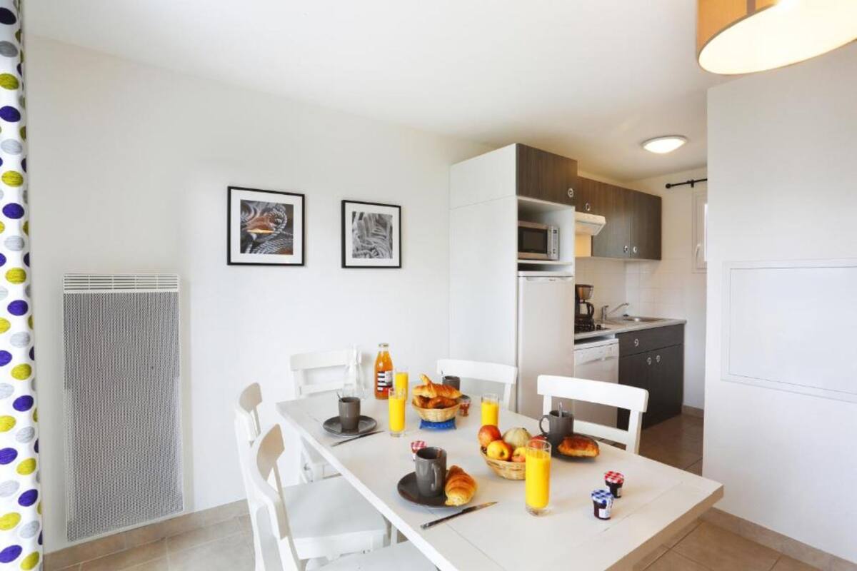 A modern kitchen with white cabinets, a microwave, and a sink. There is a dining table with chairs, a fruit bowl, and breakfast items.