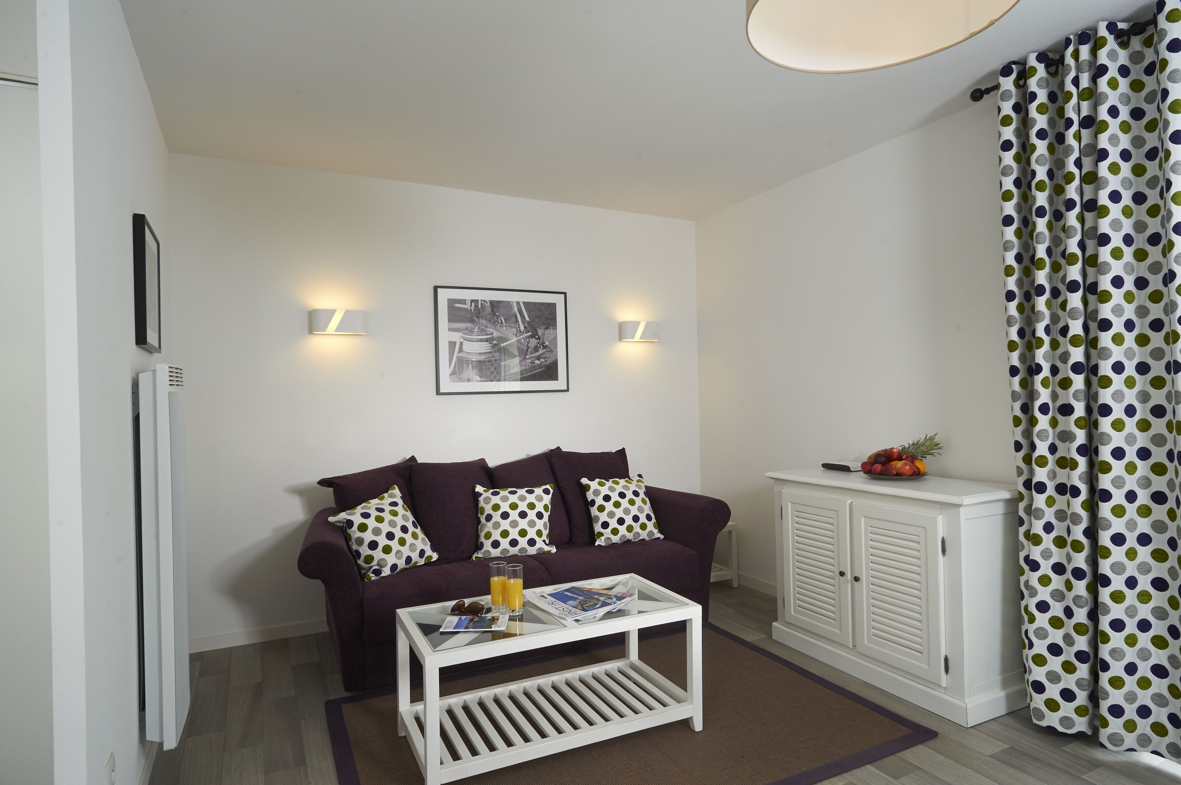A living room with a purple sofa, a white coffee table, and a white cabinet. There are two wall-mounted lights and a framed picture on the wall.