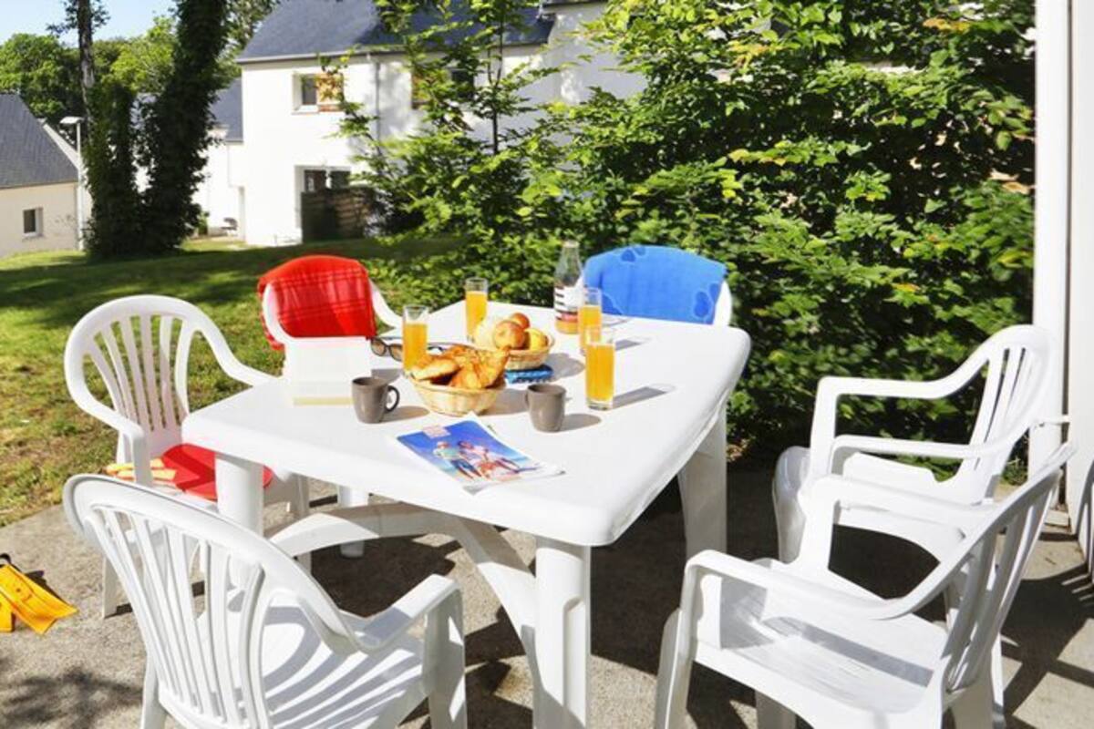 An outdoor patio with a table set for breakfast, surrounded by white chairs, in a residential area with houses and greenery.