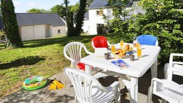 An outdoor patio with a table set for breakfast, surrounded by white chairs, in a residential area with houses and greenery.
