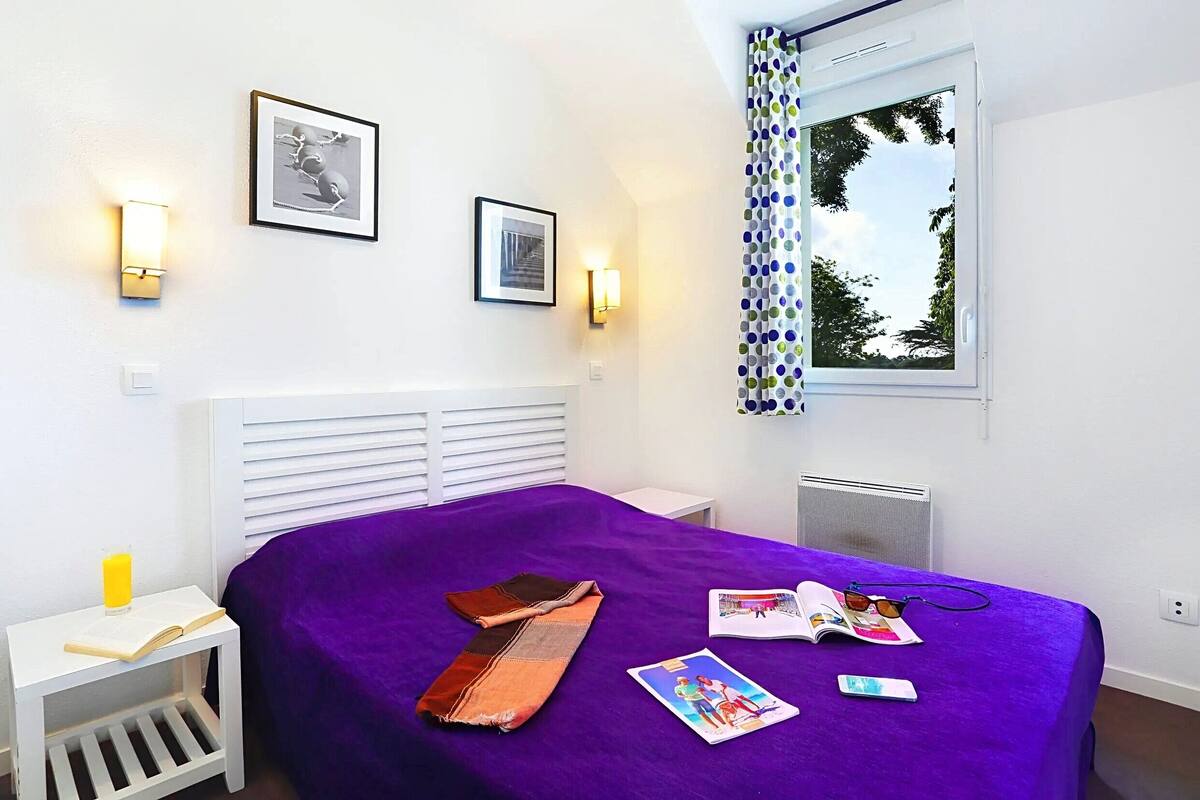A bedroom with a purple bedspread, two framed pictures on the wall, a window with a curtain, and a small bedside table with a book and a glass of orange juice.