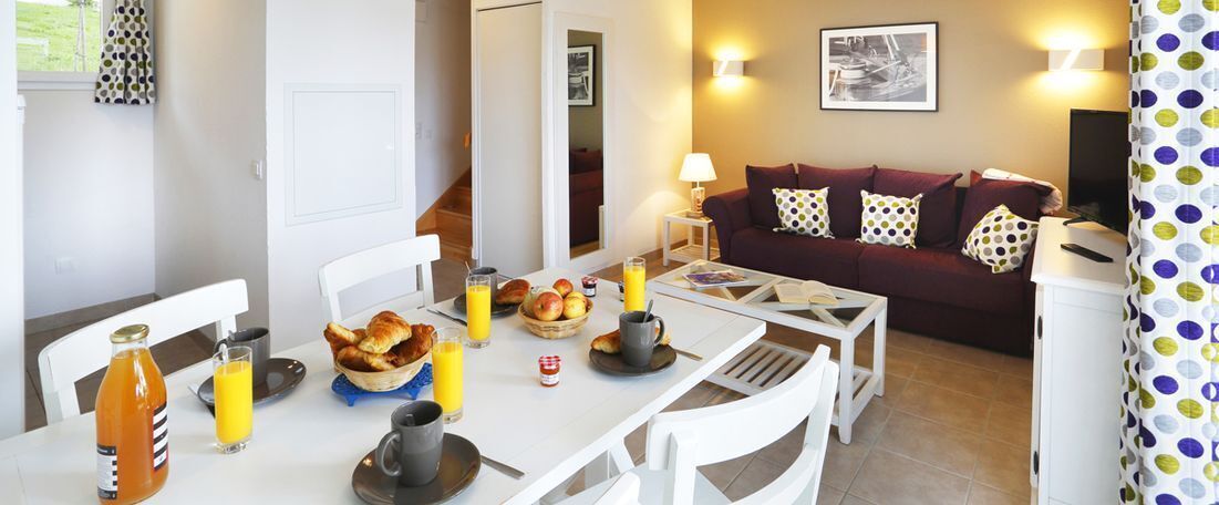 Un comedor con una mesa lista para el desayuno, incluyendo cruasanes, fruta y jugo de naranja. Se ve un sofá y un televisor al fondo.