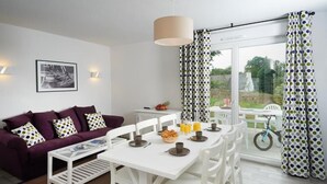 A modern living room with a purple sofa, white dining table, and a large window with curtains. - Terres de France - Le Domaine de la Baie (Audierne)