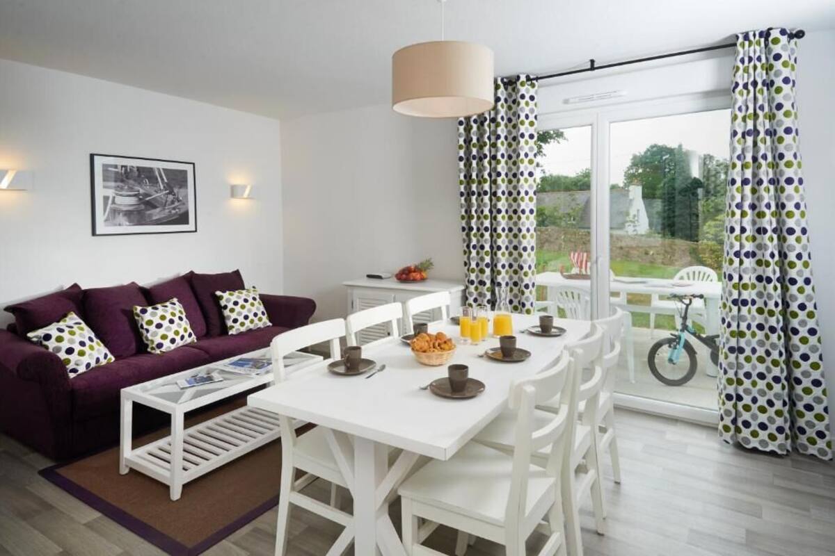 A modern living room with a purple sofa, white dining table, and a large window with curtains.
