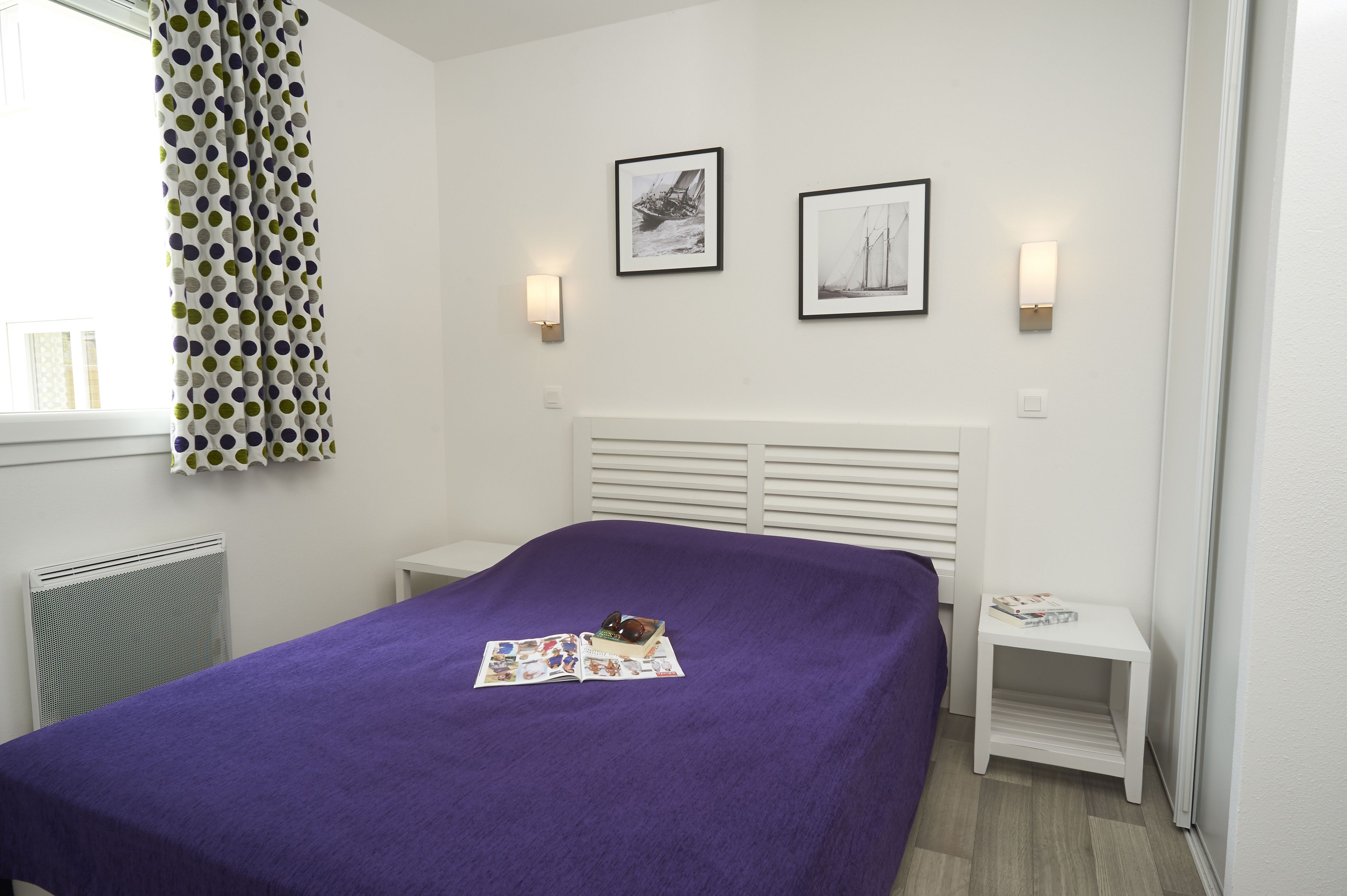 Ein Schlafzimmer mit violettem Bettbezug, einem weißen Kopfteil, zwei gerahmten Bildern an der Wand, einem weißen Nachttisch mit einem Buch und einem Fenster mit Vorhang.