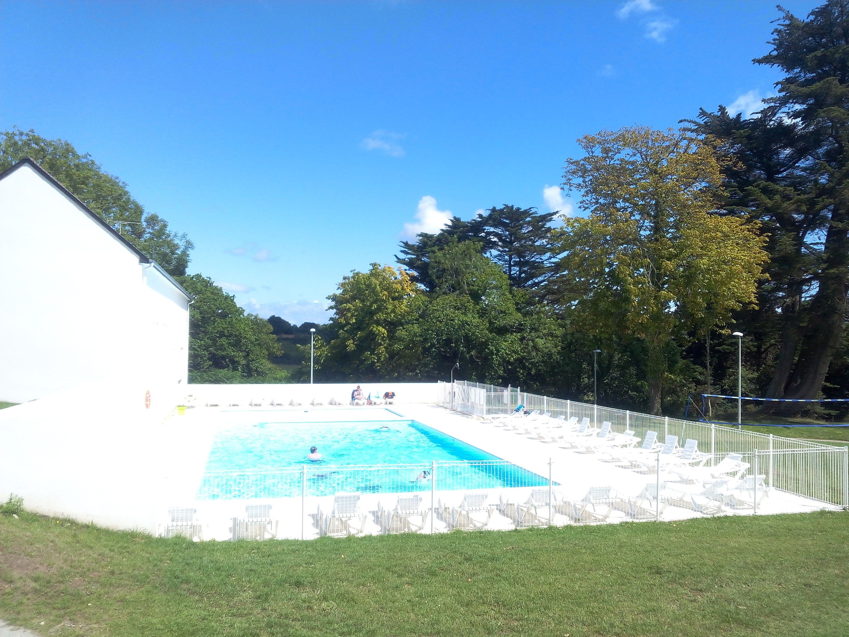 A rectangular swimming pool with a white fence and lounge chairs around it, surrounded by trees and a white building.
