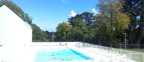 A rectangular swimming pool with a white fence and lounge chairs around it, surrounded by trees and a white building.